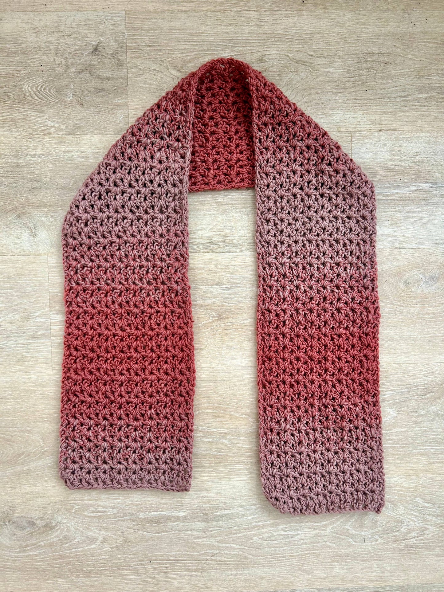 A handmade chunky crochet scarf with a gradient from light pinkish-brown to reddish-brown, laid flat on a wooden surface.