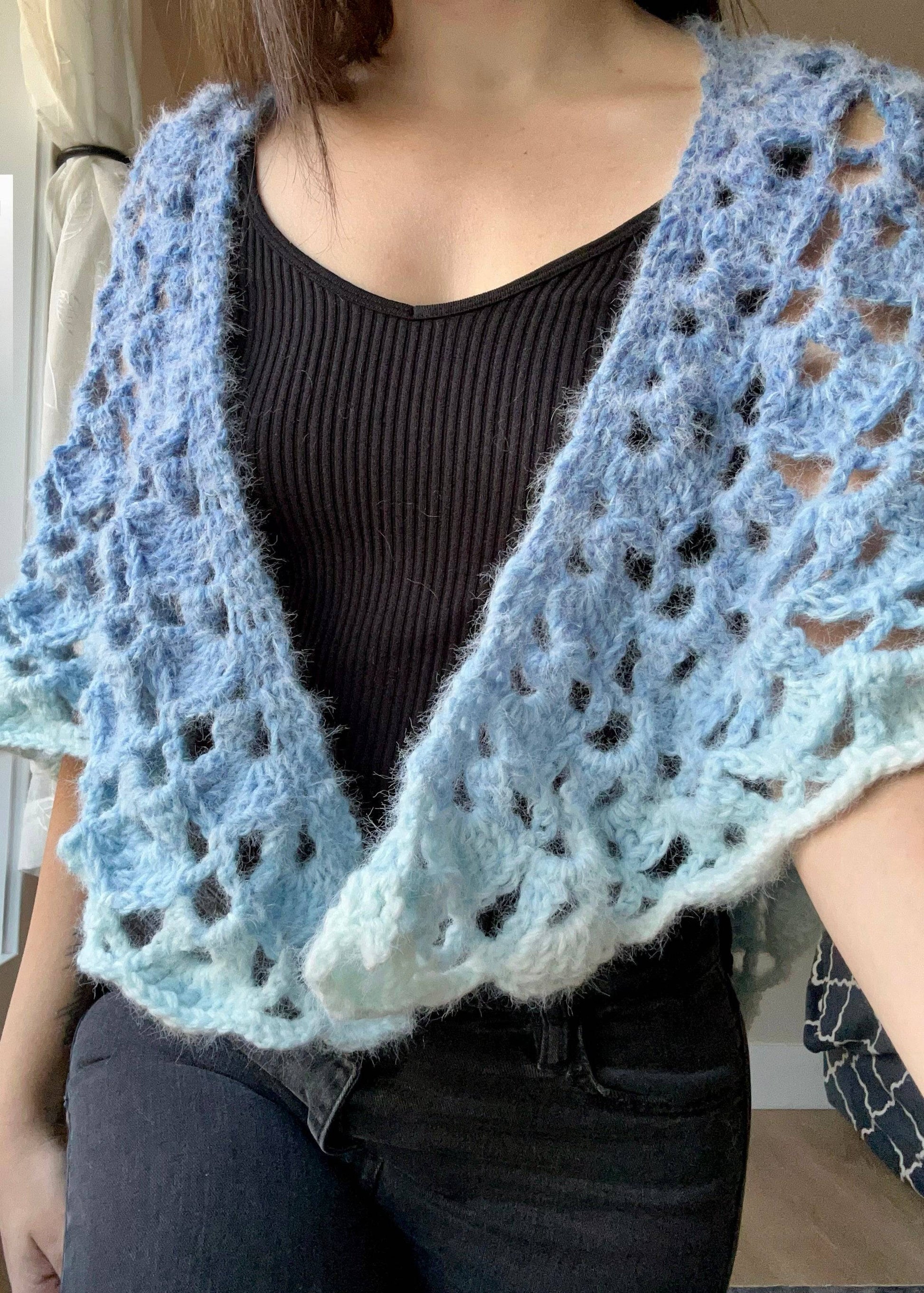 Person wearing a light blue crocheted cozy ombre shawl over a black top.