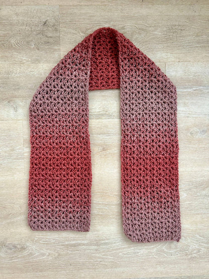 A handmade chunky crochet scarf with a gradient from light pinkish-brown to reddish-brown, laid flat on a wooden surface.